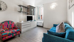 A living room with a sofa, a colorful armchair, a TV on a stand, a white cabinet, and a clock on the wall. - Garibaldi - Loano Centre Apartment for 4, Loano, Italy (Loano)