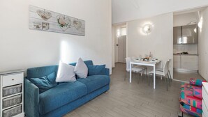 A modern living room with a blue sofa, white dining table, and a kitchen area in the background. - Garibaldi - Loano Centre Apartment for 4, Loano, Italy (Loano)