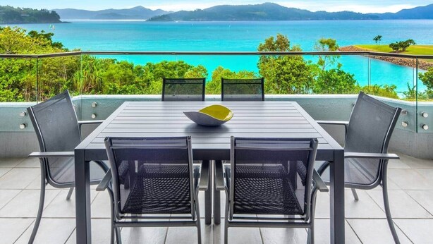 A dining area with a table and chairs, overlooking a lake with mountains in the background. - Frangipani Lodge 101 (Hamilton Island)