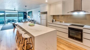 A modern kitchen with a marble island, wooden stools, a built-in oven, and a view of the outdoors. - Frangipani Lodge 101 (Hamilton Island)