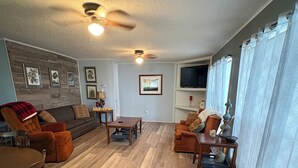 Una sala de estar con piso de madera, ventilador de techo, televisor y chimenea.