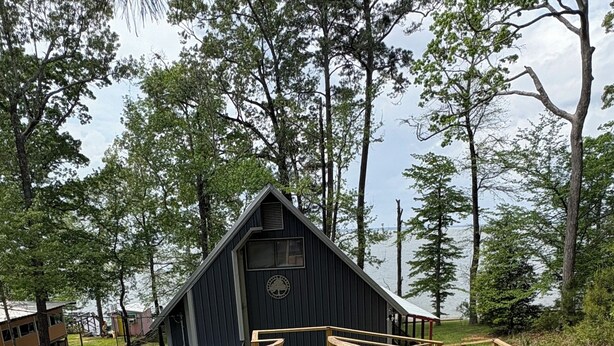 Una terraza de madera que conduce a una cabaña con techo triangular, rodeada de árboles y un cuerpo de agua.