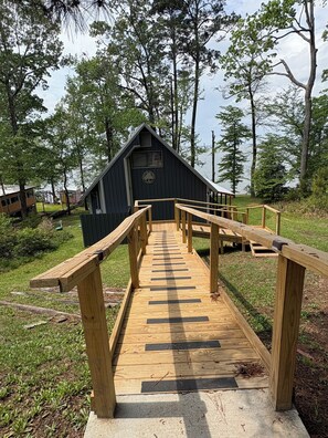 A wooden deck leading to a cabin with a triangular roof, surrounded by trees and a body of water.