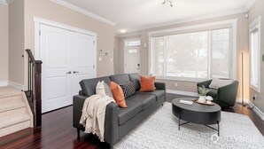 Ein Wohnzimmer mit einem dunkelgrauen Sofa, einem runden Couchtisch und einem grünen Sessel. Eine Treppe befindet sich links und ein großes Fenster rechts.