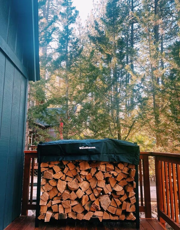 A covered stack of firewood on a wooden deck with a view of tall trees in the background.