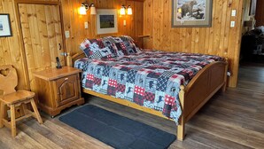 A wooden-paneled room with a bed, a nightstand, a chair, and a framed picture on the wall.