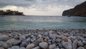 Una playa de guijarros con aguas tranquilas, una costa rocosa a lo lejos y un edificio de varios pisos.