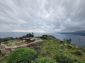 A secluded outdoor seating area with two lounge chairs and a wooden table, overlooking a coastal landscape with a view of the sea and distant hills.