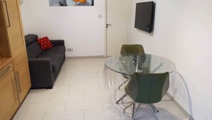 Um espaço de estar pequeno e moderno, com mesa e cadeiras de vidro, sofá cinza com almofada vermelha, e uma porta com uma imagem de uma flor na parede.