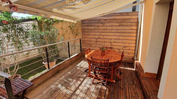Um deck de madeira com uma mesa redonda e cadeiras, rodeado por plantas em vasos e um banco de metal.