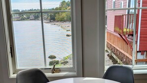 Ein Zimmer mit einem runden Tisch und vier Stühlen, einem Fenster mit Blick auf einen Fluss und ein rotes Gebäude, sowie einer Tür mit Griff.