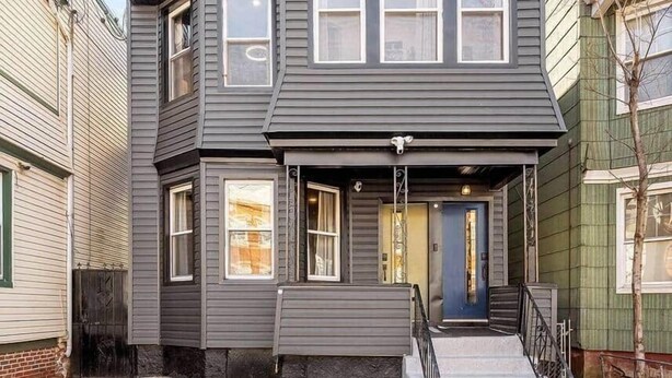 A two-story house with a gabled roof, a front porch with a black railing, and a blue door. - Cheerful Rainbow Suite • Near NYC (Jersey City)