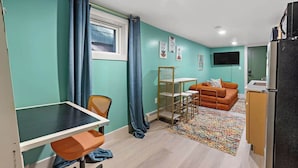 A compact living space with teal walls, a small kitchenette, a sofa, a dining table, and a TV mounted on the wall. - Cheerful Rainbow Suite • Near NYC (Jersey City)