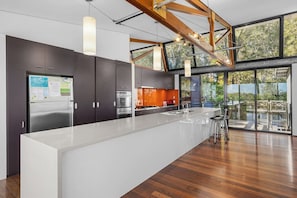 A modern kitchen with a large island, stainless steel appliances, and a view of the outdoors through large windows. - Pindimar Beach Sanctuary – Absolute Beachfront (Pindimar)