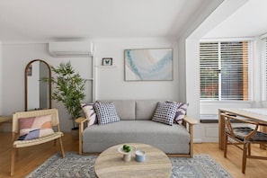 Una sala de estar moderna con un sofá gris, una mesa redonda de centro con dos objetos decorativos, un espejo grande y una planta en maceta.