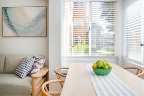 Una sala de estar acogedora con un sofá, una mesa de comedor con un tazón de fruta y un ventanal con persianas.