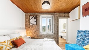 Ein Schlafzimmer mit Steinwand, einem Bett mit weißer Bettwäsche, einer Holzbalkendecke und Blick auf ein Gebäude.