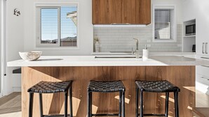 Una cocina moderna con armarios blancos, una isla de madera con taburetes y ventanas amplias con persianas.