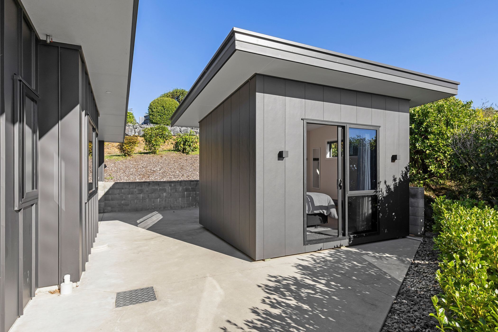 A modern building with a covered outdoor area, a door leading to a room, and a small garden area with shrubs.