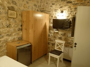 A compact room with a stone wall, a wooden wardrobe, a small fridge, a white chair, and a television mounted on the wall. - Torre Agetria Episkopē (Stavri)
