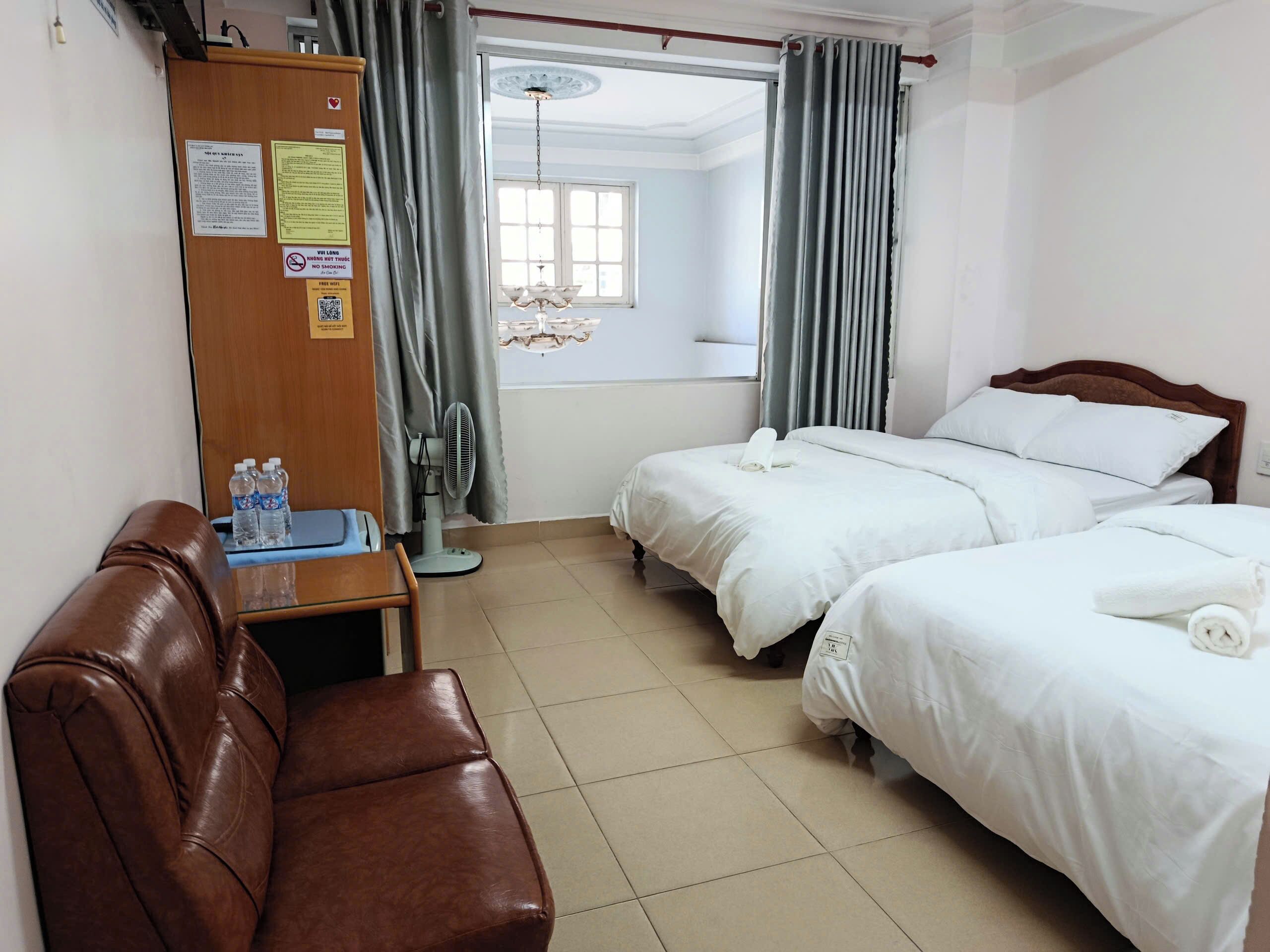 A hotel room with two beds, a wooden wardrobe, a desk with a chair, a fan, and a view of the outside.