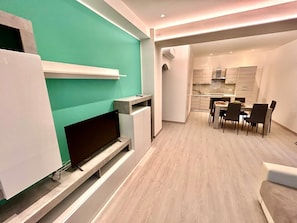 Ein modernes Wohnzimmer mit Flachbildfernseher, einem Essbereich mit Tisch und Stühlen sowie einer Küche im Hintergrund.