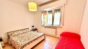 Een slaapkamer met een bed, een rode stoel, een raam met gordijnen, een radiator en een hanglamp.