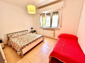 A bedroom with a bed, a red chair, a window with curtains, a radiator, and a hanging light fixture. - Trilo Roberto - Marcelli (Marcelli)