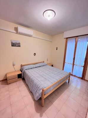 Ein Schlafzimmer mit einem Einzelbett, Nachttischen, einer Deckenleuchte, einer Klimaanlage, einem Fenster mit Vorhängen und einem kleinen Bild an der Wand.