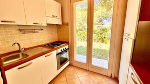 A compact kitchen with white cabinets, a gas stove, an oven, and a sink. There is a sliding glass door leading to a garden. - Two-room apartment Leonardo - Sirolo (Sirolo)