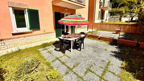A patio with a table and chairs, a colorful striped umbrella, and a building with green shutters and a balcony. - Two-room apartment Leonardo - Sirolo (Sirolo)