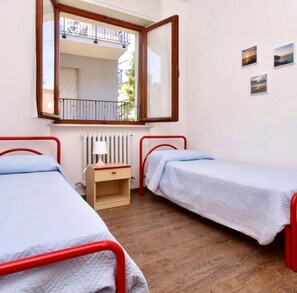 A room with two single beds, a small wooden nightstand, a radiator, and a window with a view of the outside balcony. - TRILO LARA with wifi and parking (Sirolo)