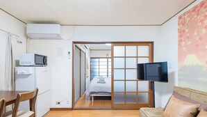 Ein kleines Zimmer mit einem Bett, einem an der Wand befestigten Fernseher, einer Mikrowelle und einem Essbereich mit Tisch und Stühlen.