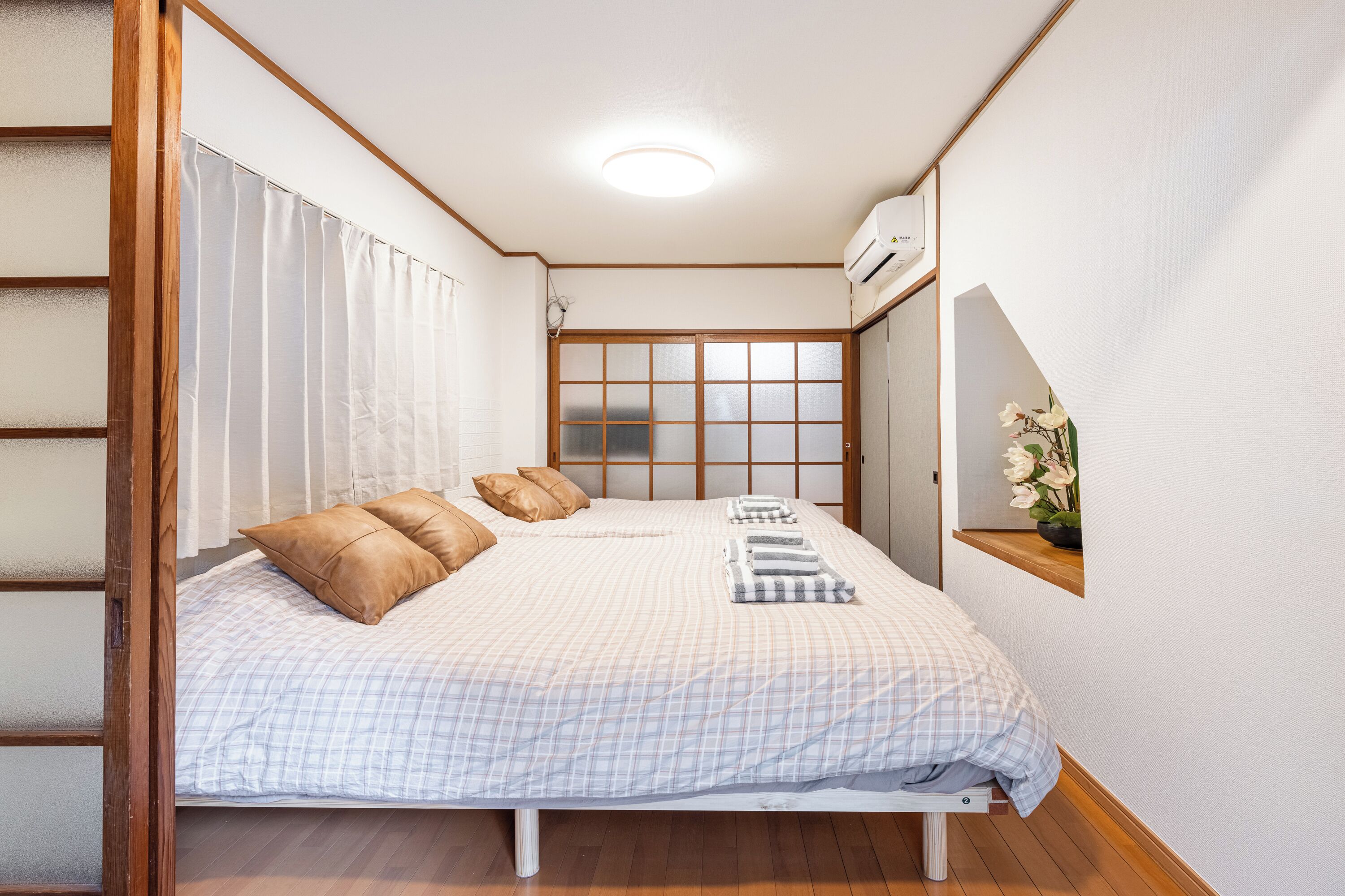 Een slaapkamer met een bed, kussens, een nachtkastje, een raam met jaloezieën en een klein plankje met een plant.
