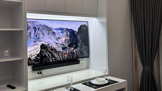 Ett modernt rum med en stor TV med platt skärm som är monterad på ett vitt skåp, ett litet bord med två glas och en skål samt ett par stolar.
