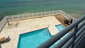 Área de piscina com vista para o mar, um deck com cadeiras e um corrimão branco.