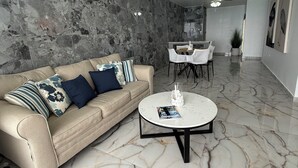 Uma sala de estar moderna com piso de mármore, um sofá bege com almofadas decorativas, uma mesa de centro redonda e um ventilador de teto.