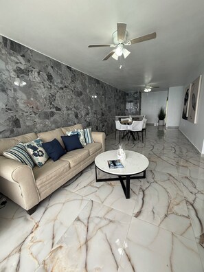 Una sala de estar moderna con piso de mármol, un sofá color beige con cojines decorativos, una mesa de centro redonda y un ventilador de techo.