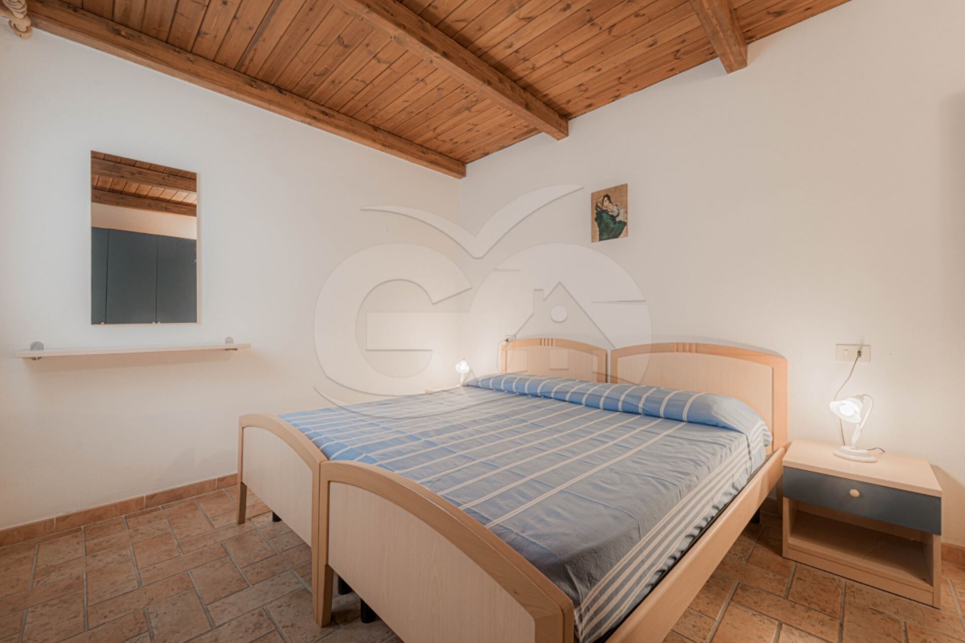 A bedroom with a wooden bed, a nightstand, a wall-mounted shelf, and a ceiling with exposed beams.