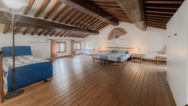Un amplio dormitorio en el ático con vigas de madera, una cama grande, una cama más pequeña, un ventilador y un escritorio.