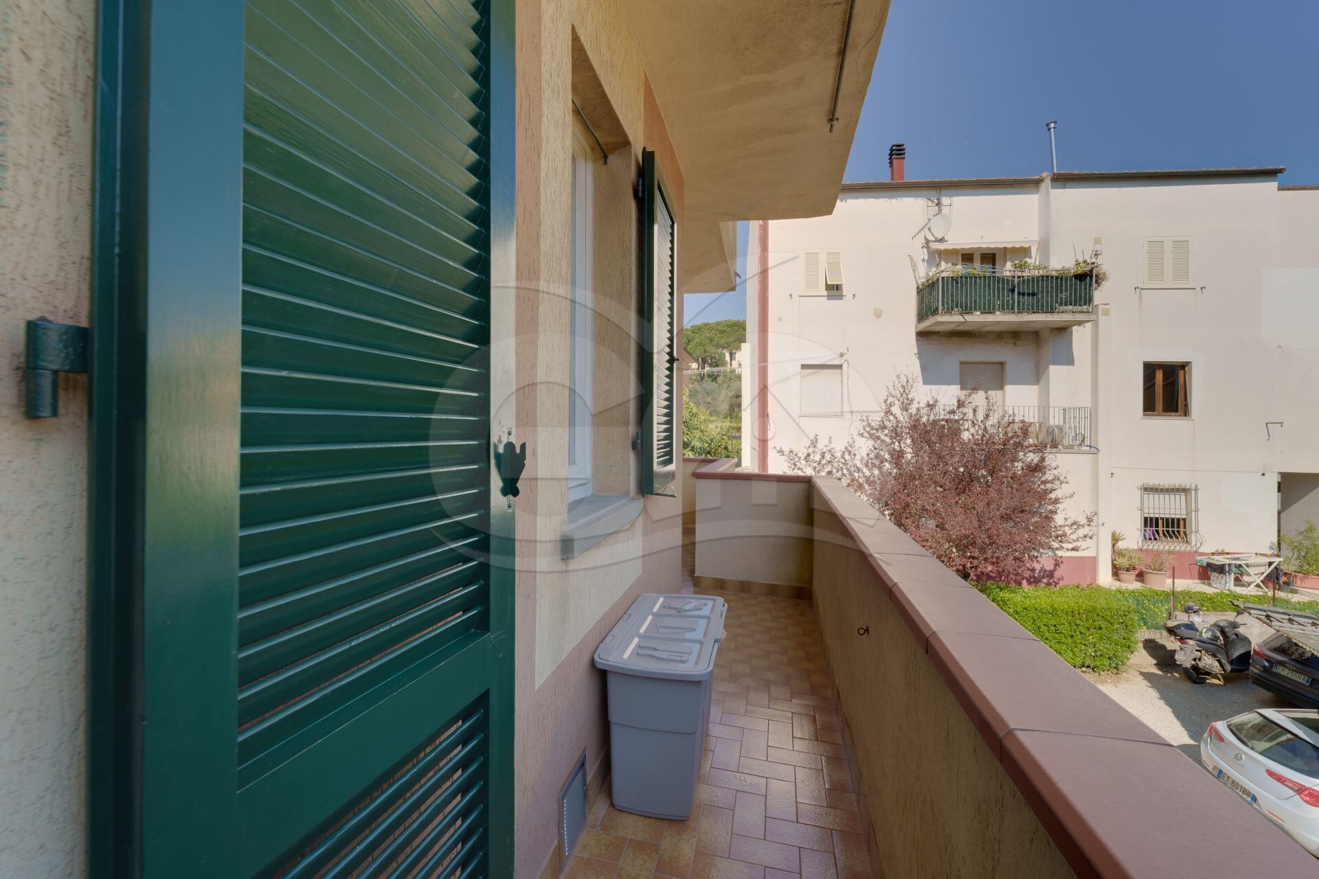 A balcony with a trash bin, a clear view of the street, and a building with a balcony in the background.