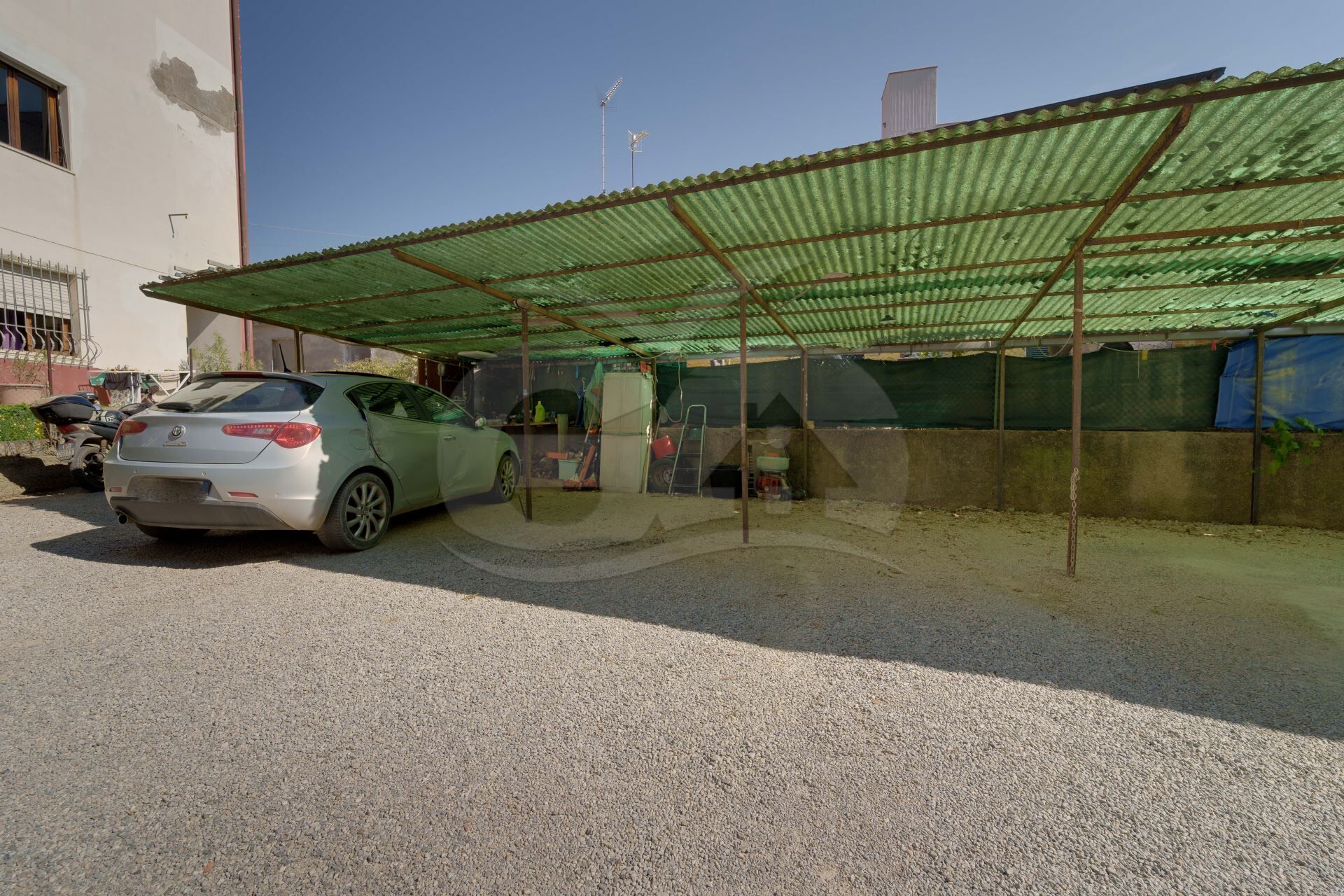 Un'auto parcheggiata sotto una struttura verde in metallo, un edificio con facciata bianca e un cielo azzurro intenso.