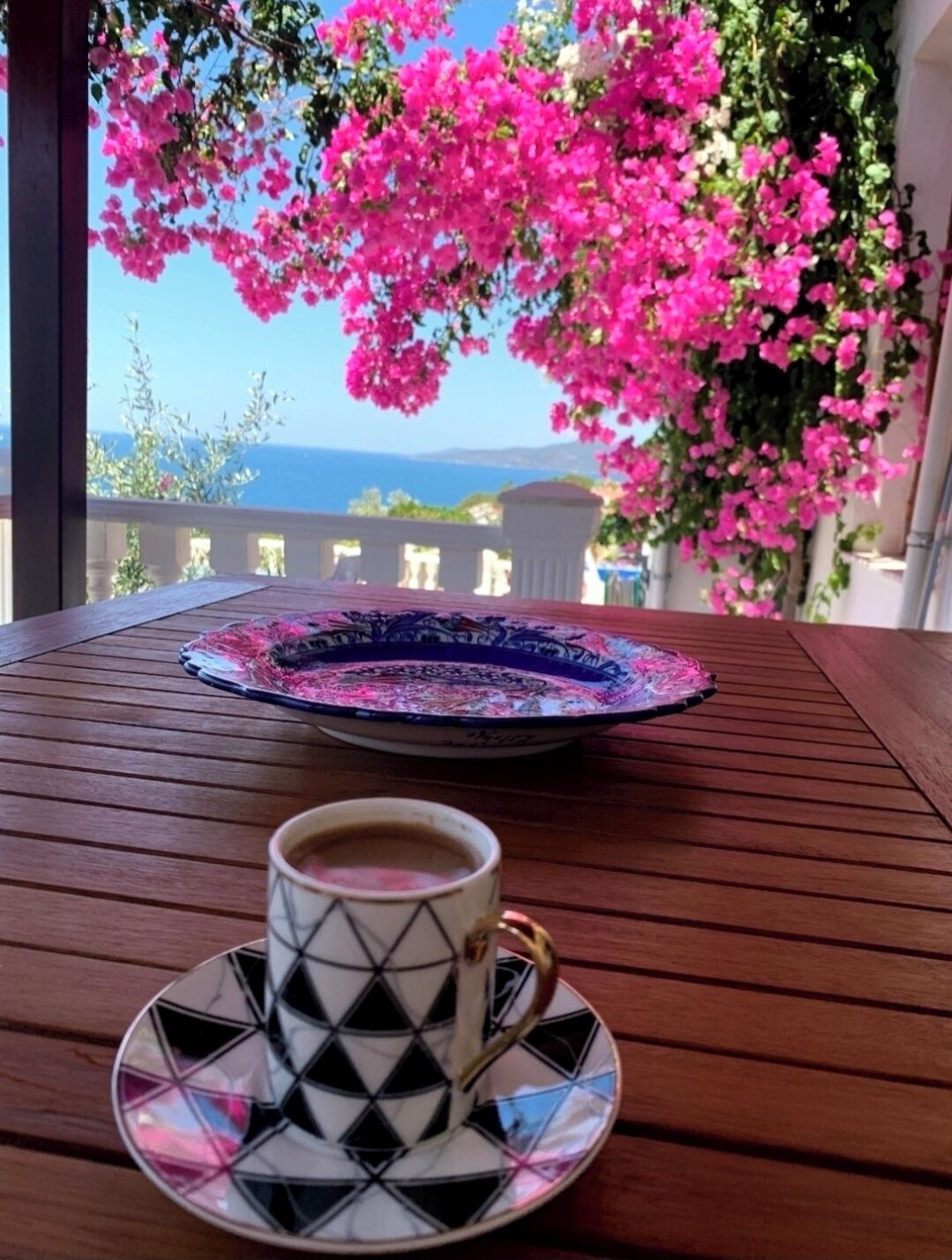 A table with a cup of coffee and a decorative plate, set against a backdrop of pink flowers and a view of the sea.