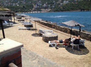 Zona de playa con sillas de descanso y sombrillas, un muro de piedra, y vistas al mar y casas en la colina.