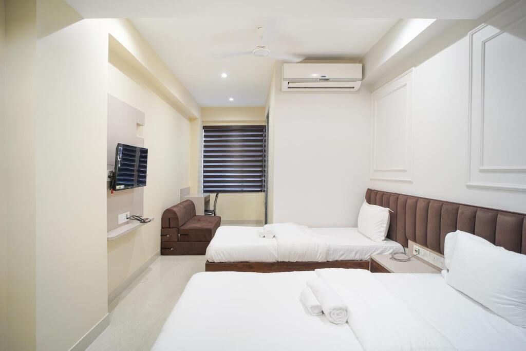 A hotel room with two beds, a sofa, a ceiling fan, an air conditioning unit, and a window with blinds.