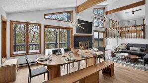 A spacious living area with a large dining table, a fireplace, and multiple windows offering views of the outdoors. - Luxury 5BR Poconos Retreat | Ski, Lake & Resort (Saw Creek)