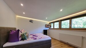 Ein Schlafzimmer mit einem Bett, einem an der Wand montierten Fernseher, Holzboden, einem Fenster mit Blick auf Bäume und einem Heizkörper.