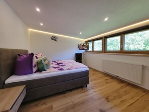 Ein Schlafzimmer mit einem Bett, einem an der Wand montierten Fernseher, Holzboden, einem Fenster mit Blick auf Bäume und einem Heizkörper.