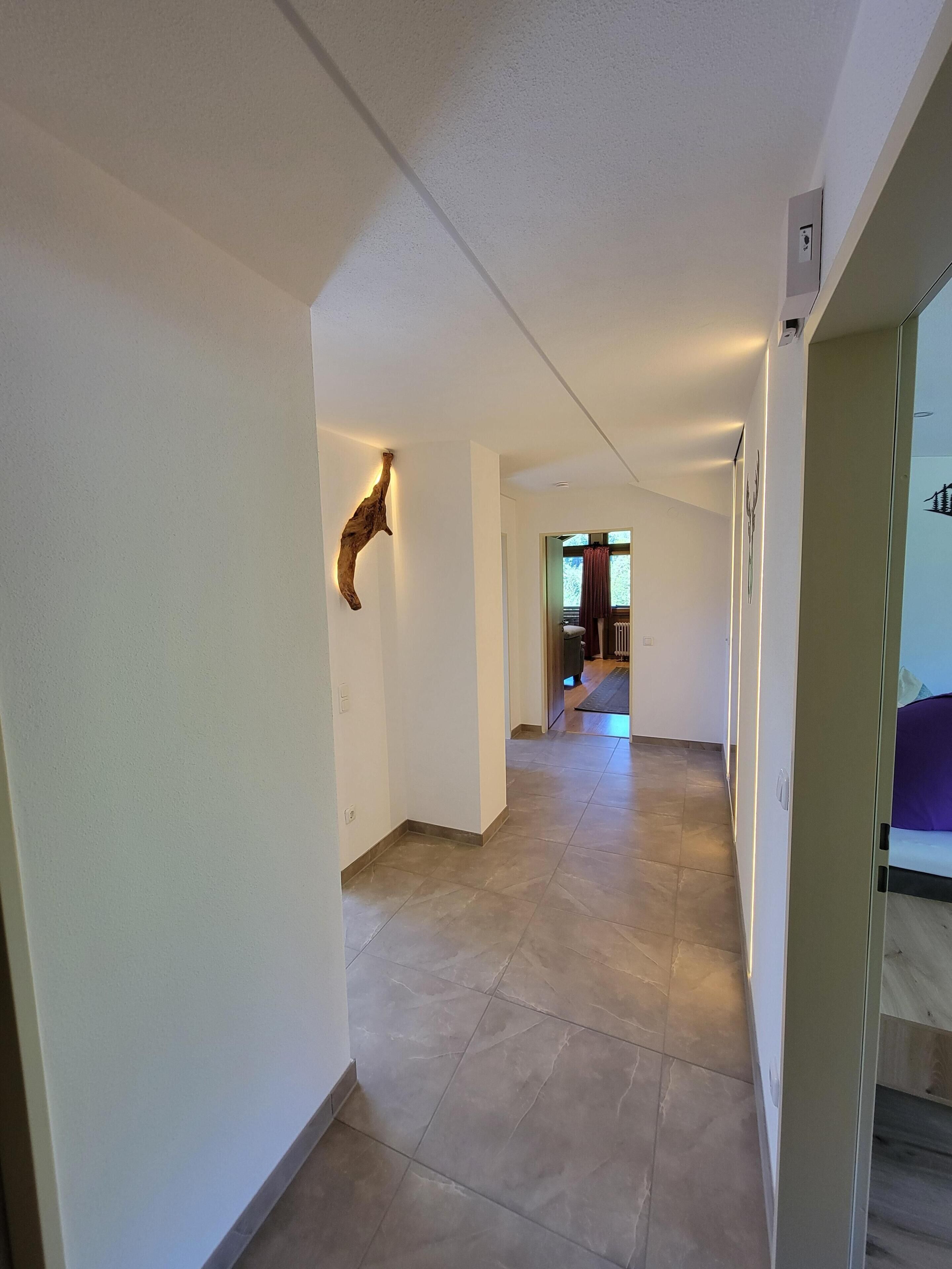A hallway with a wooden wall accent, a door leading to a room with a bed, and a view of a window.