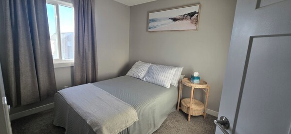 A bedroom with a bed, curtains, a bedside table with a basket, a wall-mounted picture, and a door. - Clean & Stylish 3BR Home w/ Bonus Room Near YYC, Shopping & Banff (Calgary)